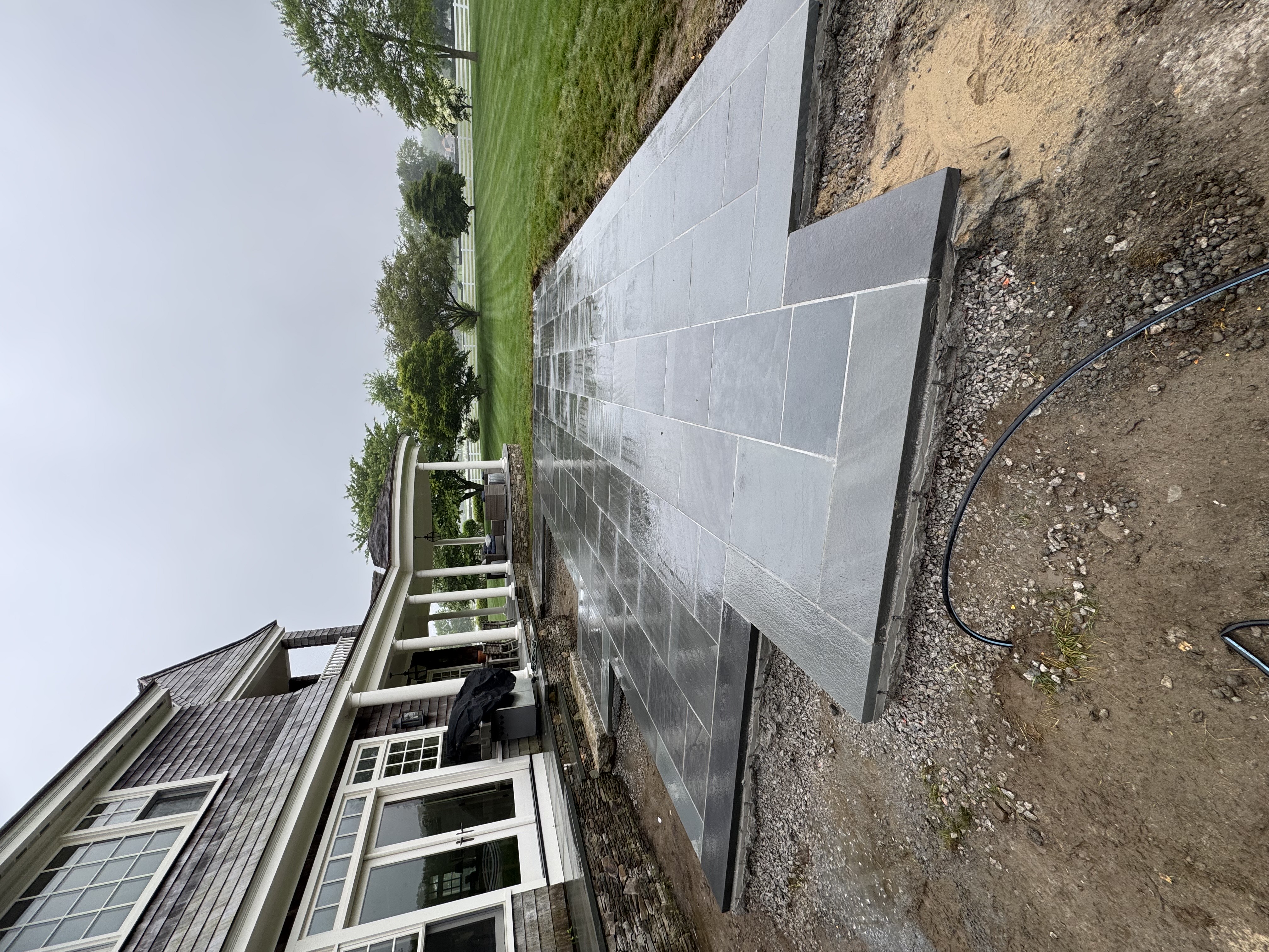 Wet stone patio under construction near shingle-sided house with white columns